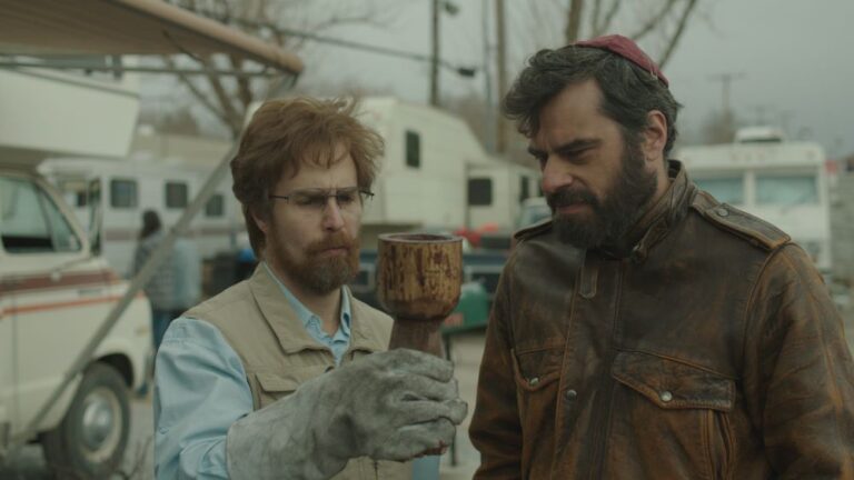 Red-bearded and bespectacled man wearing heavy gloves holds a chalice as he and another man, with dark hair and wearing a red yarmulke, peer solemnly at it. A motor home, a mini motor home, and other camping vehicles are in the background.