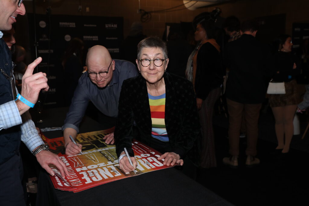 a white woman looking up at the camera at a table where she is signing a poster, a man is right behind her looking down