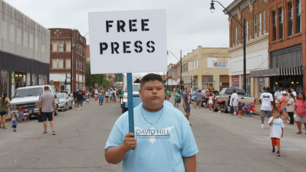 A young boy with short hair and a blue t-shirt stnads in the street with a sign that reads "Free Press"