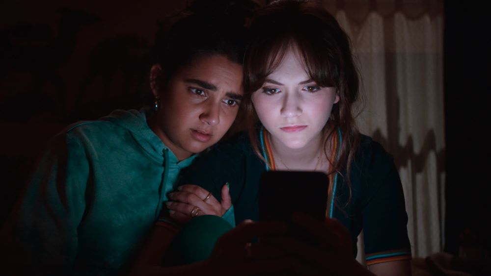 Darker-skinned young woman, with a concerned look on her face, and white young woman peer at a laptop screen in a darkened room