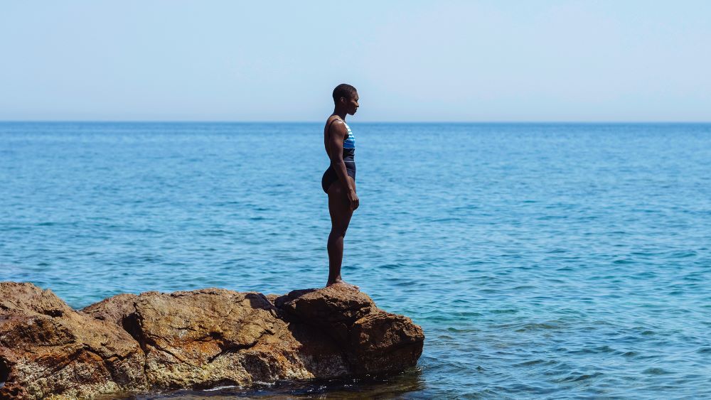 A Black woman in a bathing suit stands on an outcropping of rocks jutting into a body of water