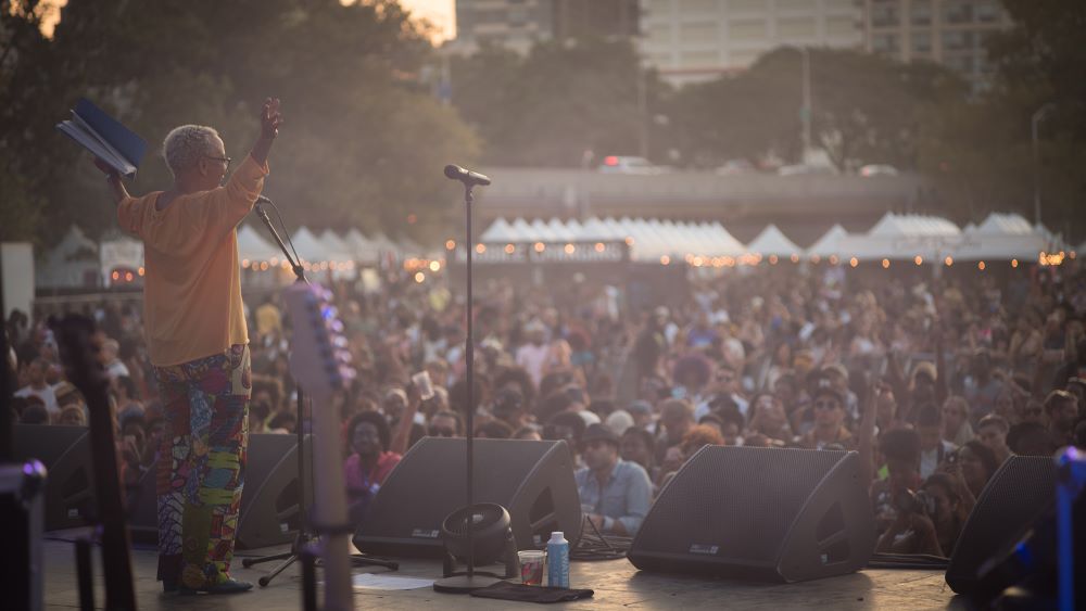 A Black woman with short gray hair stands on an outdoor stage, in a bright orange top and patterned colorful pants, with her arms upraised. A crowd stretches before her, white-peaked tents and city buildings behind them