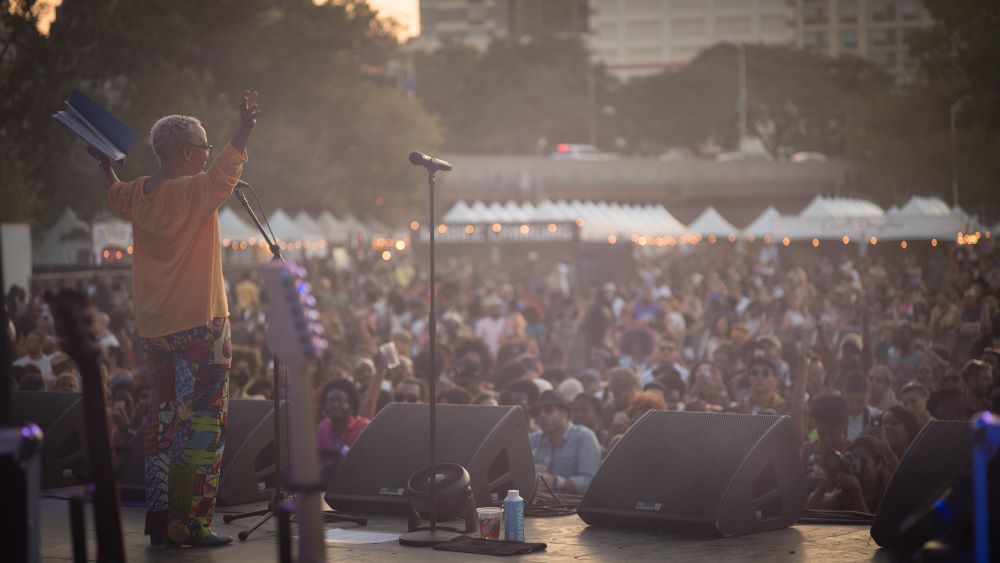 A Black woman with short gray hair stands on an outdoor stage, in a bright orange top and patterned colorful pants, with her arms upraised. A crowd stretches before her, white-peaked tents and city buildings behind them