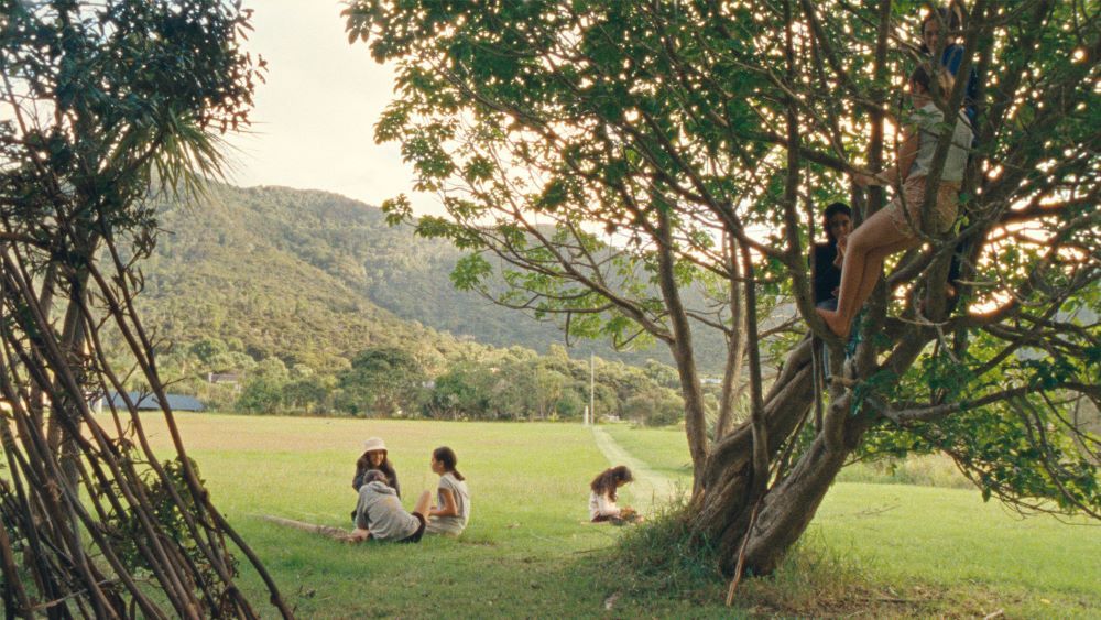 Children in summer clothing sit in the lower branches of a tree and in the lush field surrounding it. Tree-covered hills are in the background.