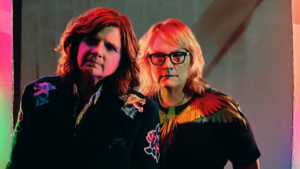 A close up of The Indigo Girls - Two woman musicians, one with brown hair and one blond with glasses. Both are wearing black and looking straight into the camera
