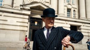 Older man in a dark bowler hat, pin-striped suit, and carrying a walking stick, checks his watch while standing in front of what appears to be a giant, white bureaucratic building. Pedestrians walk behind him.