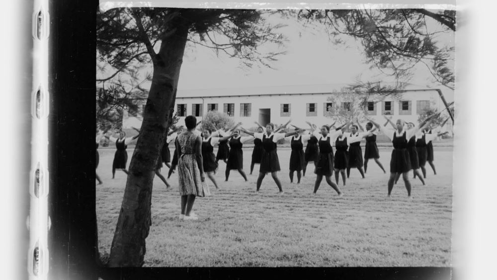 Black and white photo of what appears to be a class of Black girls in school uniforms (dark jumpers, white short-sleeve shirts) doing jumping jacks in rows, in an outdoor setting, a grassy area between a tree and a large white building. An adult Black woman stands facing them, apparently leading the exercise.