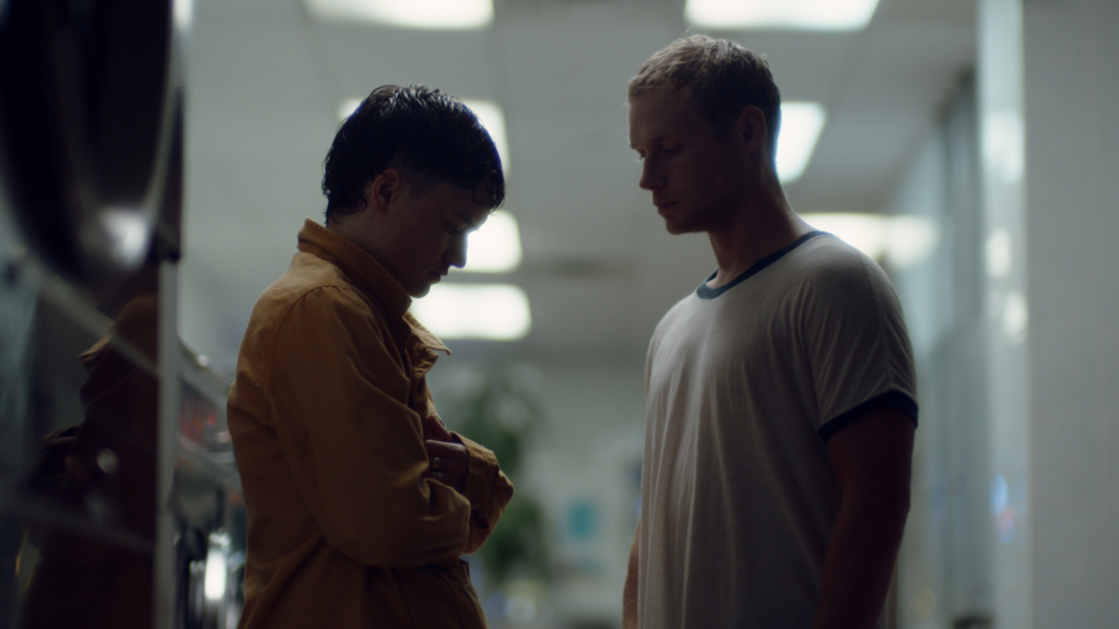 Two men - one in brown and one in a white tshirt - stand in a hallway with bright lights