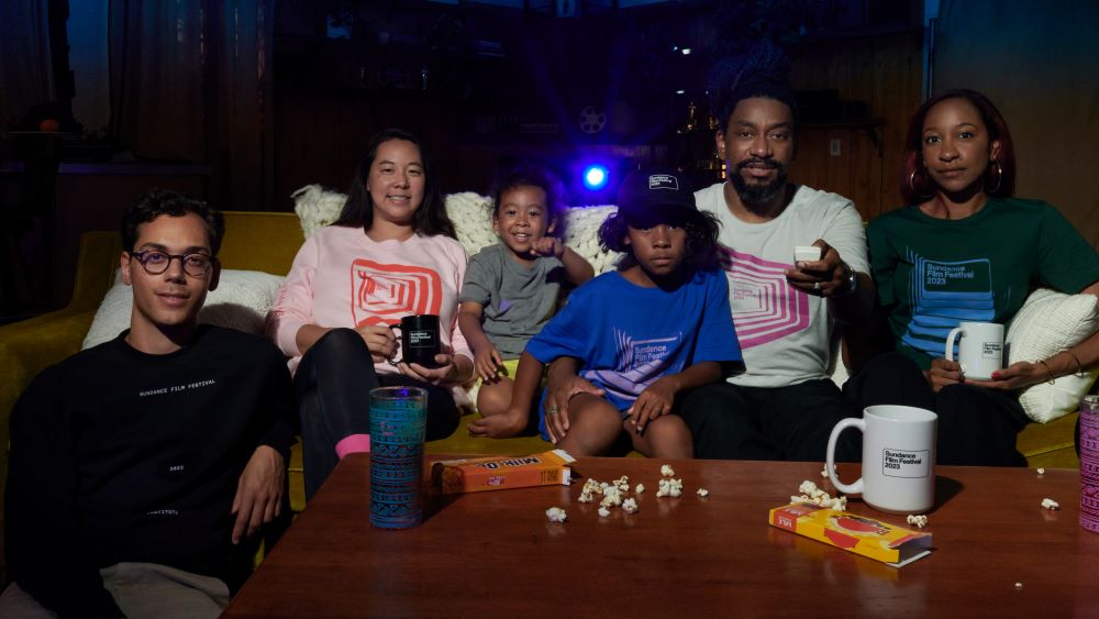 Four adults and two children are gathered on a couch and on the floor, one adult pointing a remote at the camera, which stands in for a television screen