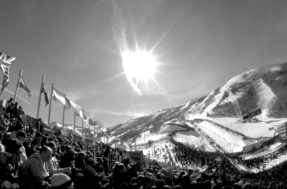 Black and white photo with the sun shining brightly on a snow-covered mountainous landscape, with various national flags flying and hoards of spectators watching what seems to be a snow-sport sporting event.