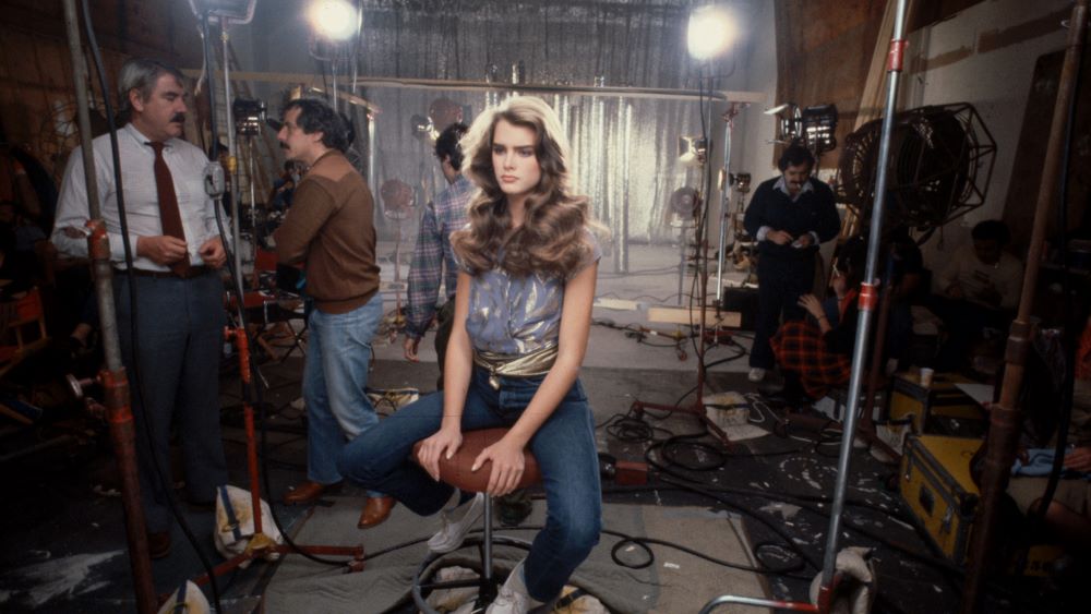 Young woman with long, flowing hair sits on a stool under lights . Men are conversing and working in the background.