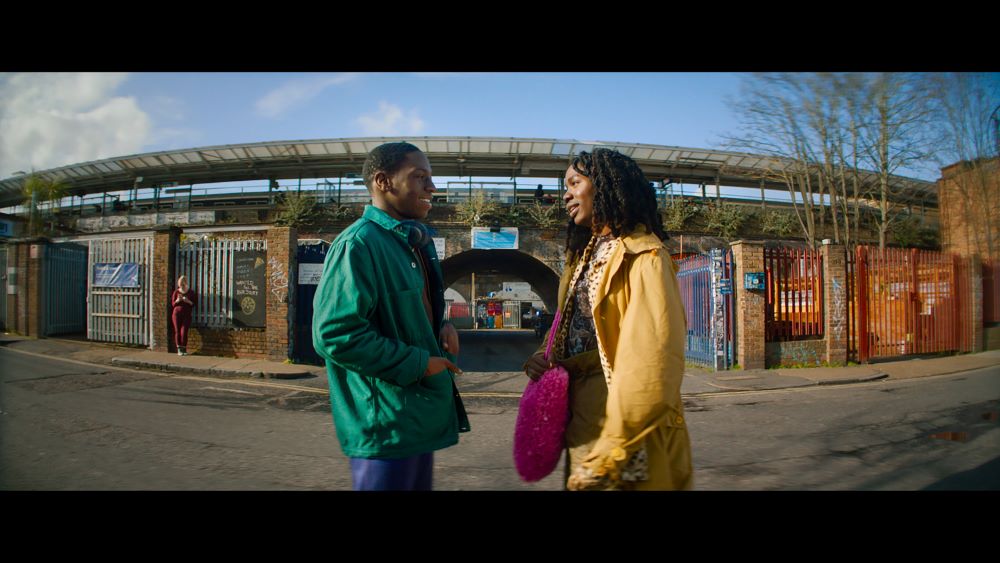 Black man in green jacket, Black woman in gold jacket, with a bright pink large purse, smiling and facing each other in front of what appears to be some kind of outdoor facility