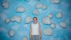 Young girl in white tank top and gray pants stands in front of a giant mural of a blue sky with fluffy white clouds, a look of awe upon her face