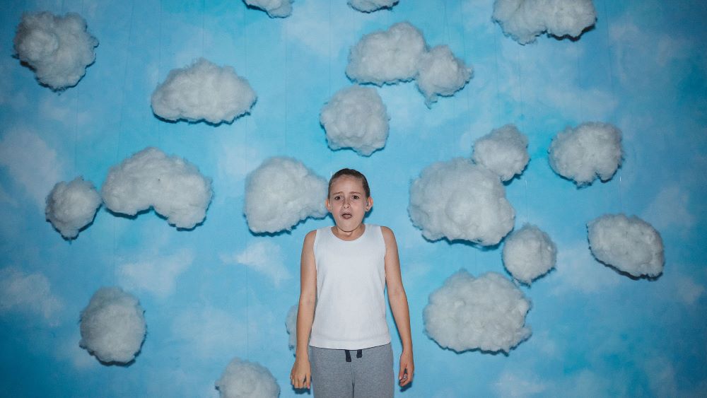Young girl in white tank top and gray pants stands in front of a giant mural of a blue sky with fluffy white clouds, a look of awe upon her face