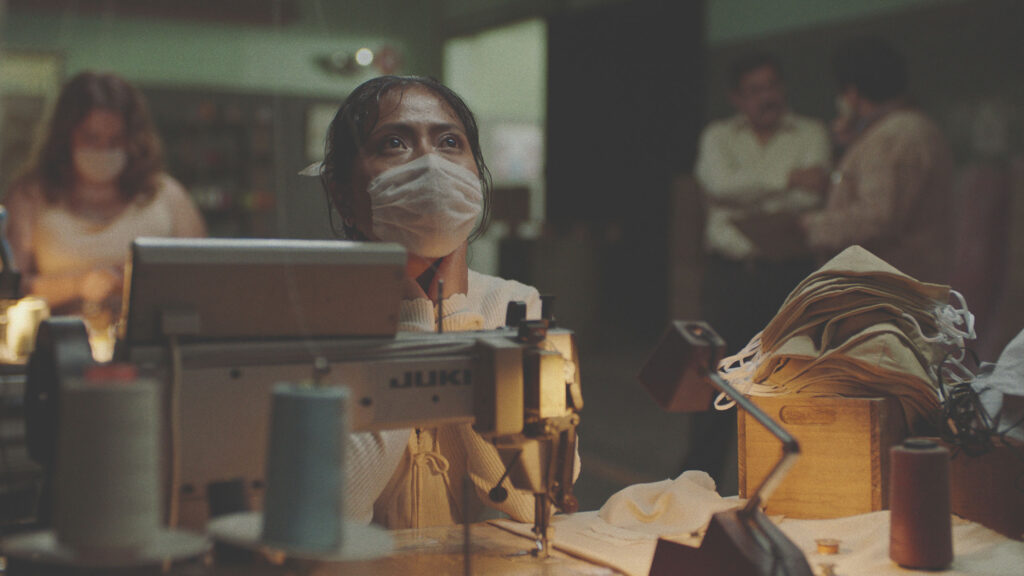 A young woman with dark hair wearing a mask looks up as she works in a sweatshop