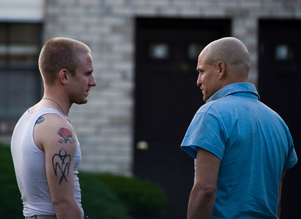 Two men stand with backs mostly to the camera, facing each other. The man on the left has closely cropped brown-red hair and tattoos on his arm and shoulder; he is wearing a white sleeveless undershirt. The man on the right looks to have a shaved head and wears a short-sleeved blue shirt.