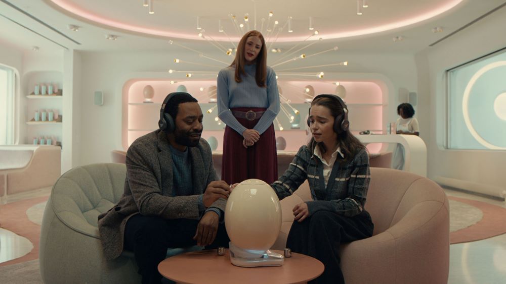 A Black man and a white woman, both wearing headphones, sit on a curved couch in a spaceship-looking room. Both are gazing at and touching a shiny white giant egg-like item on the coffee table in front of them. Behind them, a reddish-haired woman looks on.