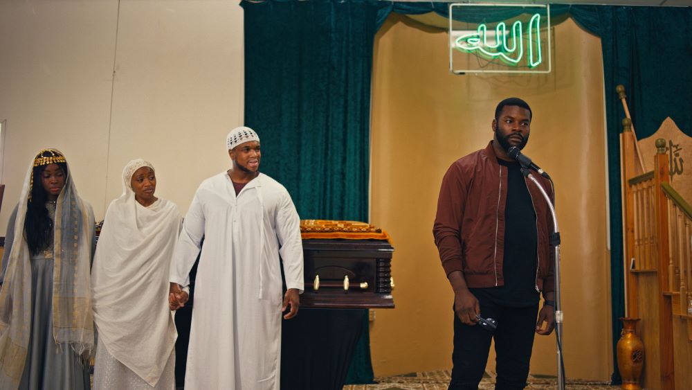 Bearded Black man stands behind a microphone and in front of a casket. To the side are two Black women and a Black man dressed in white robes