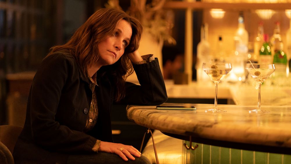 A dark-haired white woman sits at a bar, a drink in front of her, head rested on her left arm, with a despondent look on her face