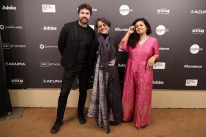 A bearded man in all black, a woman with dark grey hair and a long black scarf, and a woman in a pink dress with medium-length curly hair, all stand in front of a step and repeat at the 2023 Sundance Film Festival.