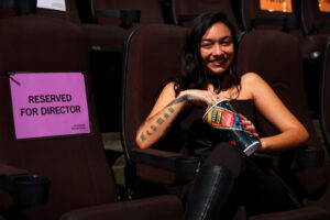 A girl with long black hair in black is eating popcorn sitting next to a sign that says, "reserved for director."