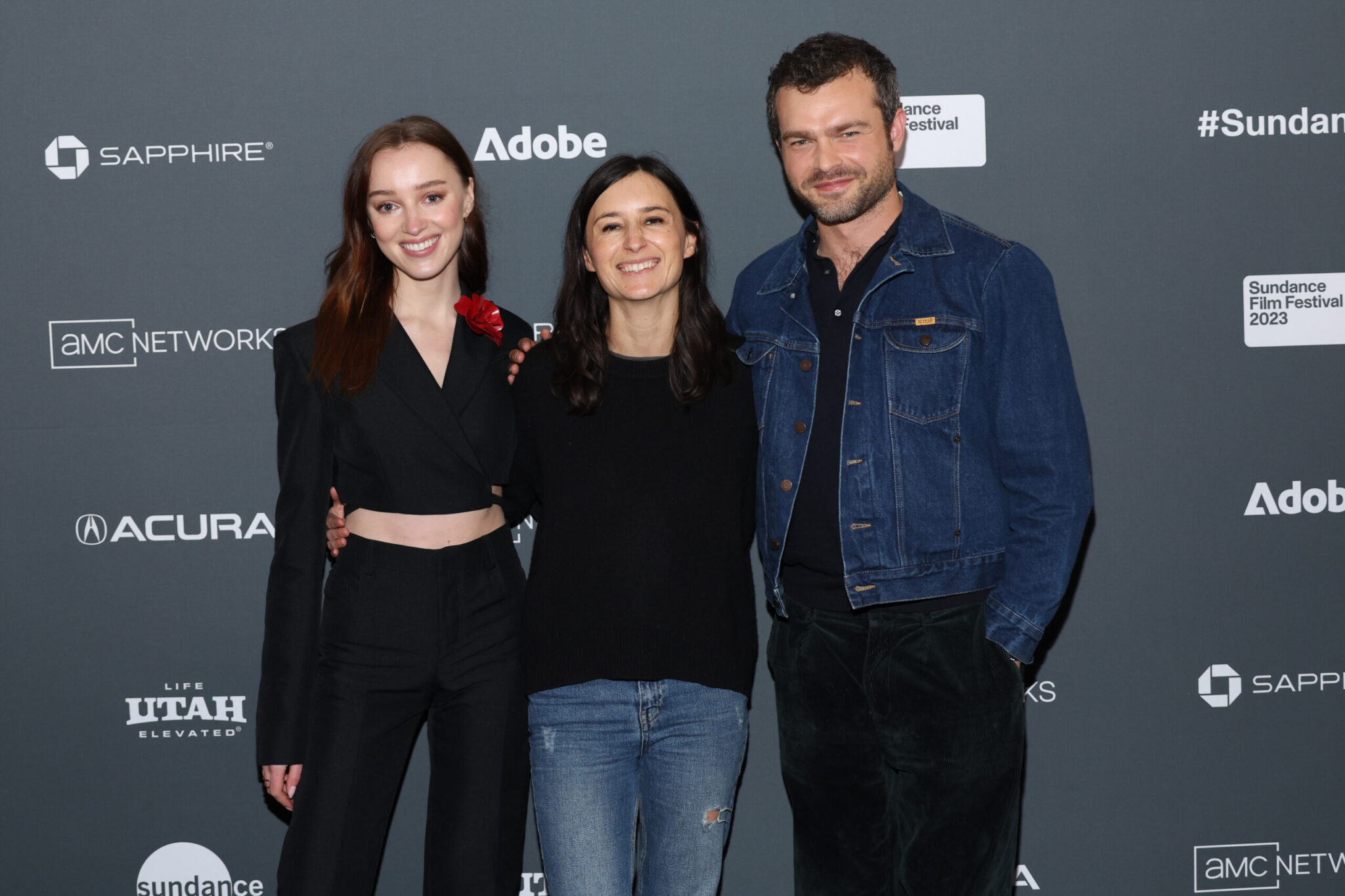 A woman with red hair dressed in all black, a woman with a black long sleeve shirt and blue jeans, and a man dressed in black with a denim jacket, stand in front of a step and repeat at the 2023 Sundance Film Festival.