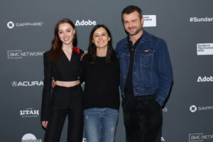 A woman with red hair dressed in all black, a woman with a black long sleeve shirt and blue jeans, and a man dressed in black with a denim jacket, stand in front of a step and repeat at the 2023 Sundance Film Festival.