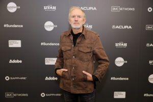 A man with grey hair in a brown coat stands in front of a step and repeat at the 2023 Sundance Film Festival.