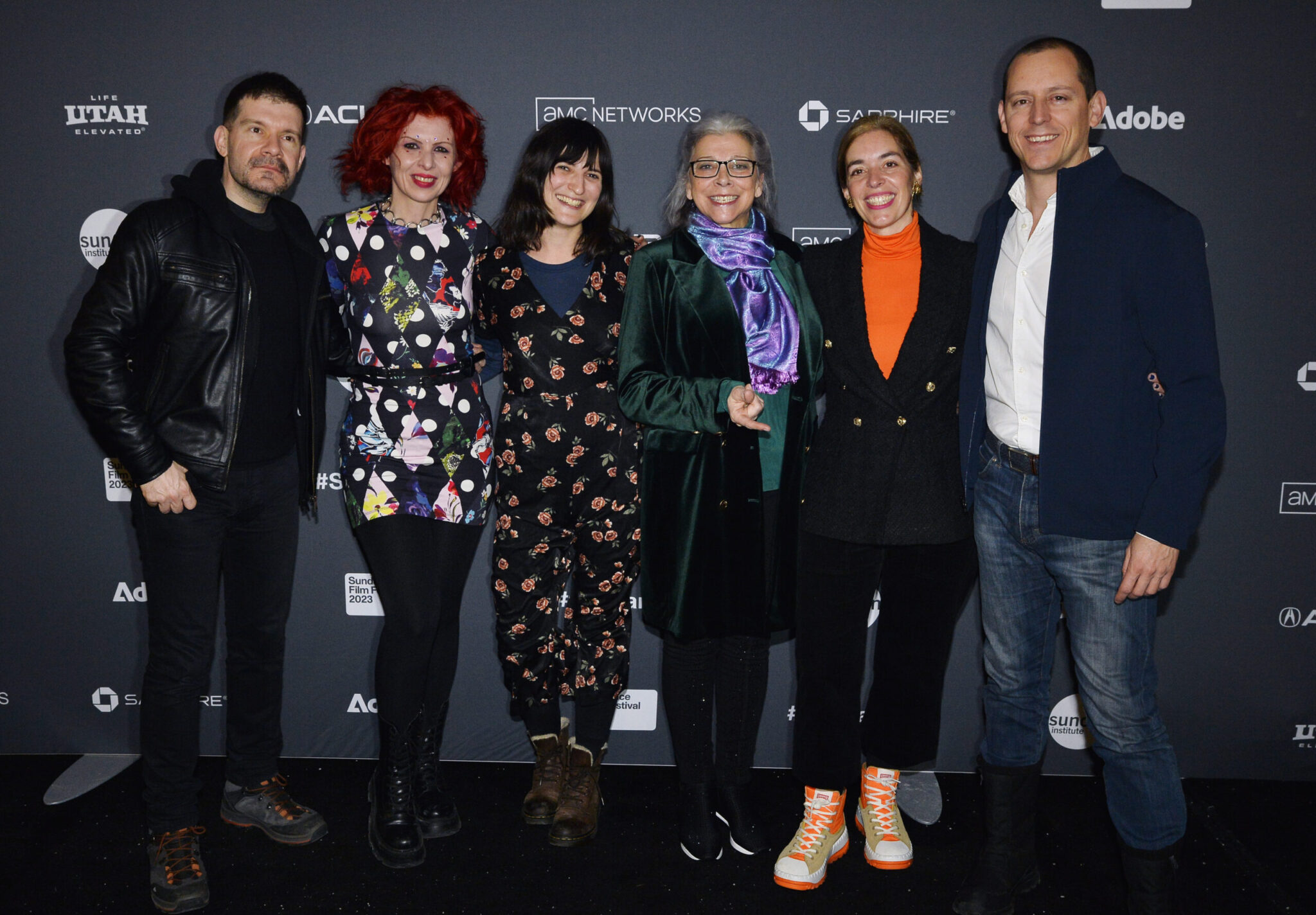 A man with dark hair in all black, a woman with red hair in a colorful dress, a woman with medium-length black hair wearing a flower dress, a woman with grey hair and a purple scarf and green coat, a woman in a black coat over an orange turtle neck, a man with a blue coat over a white shirt and jeans stand in front of a step and repeat at the 2023 Sundance Film Festival.