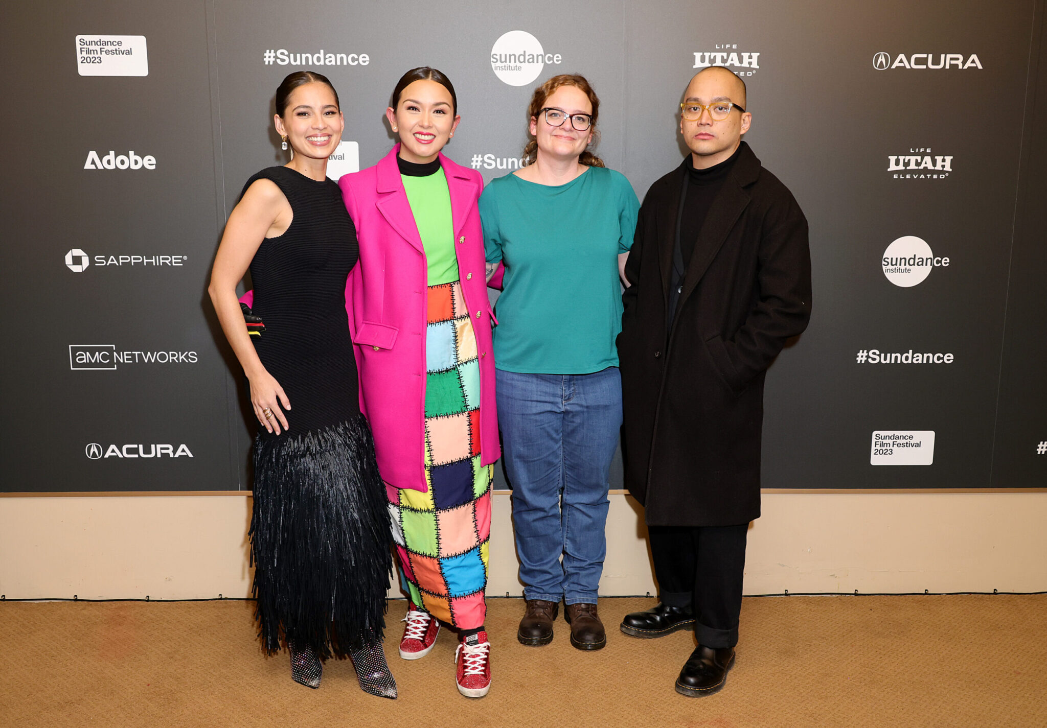 A woman with brown hair and a blakc dress, a woman with a pink coat over a colorful dress, a woman in an aqua-colored t-shirt and jeans, and a man in glasses dressed in all black, stand in front of a step and repeat at the 2023 Sundance Film Festival.