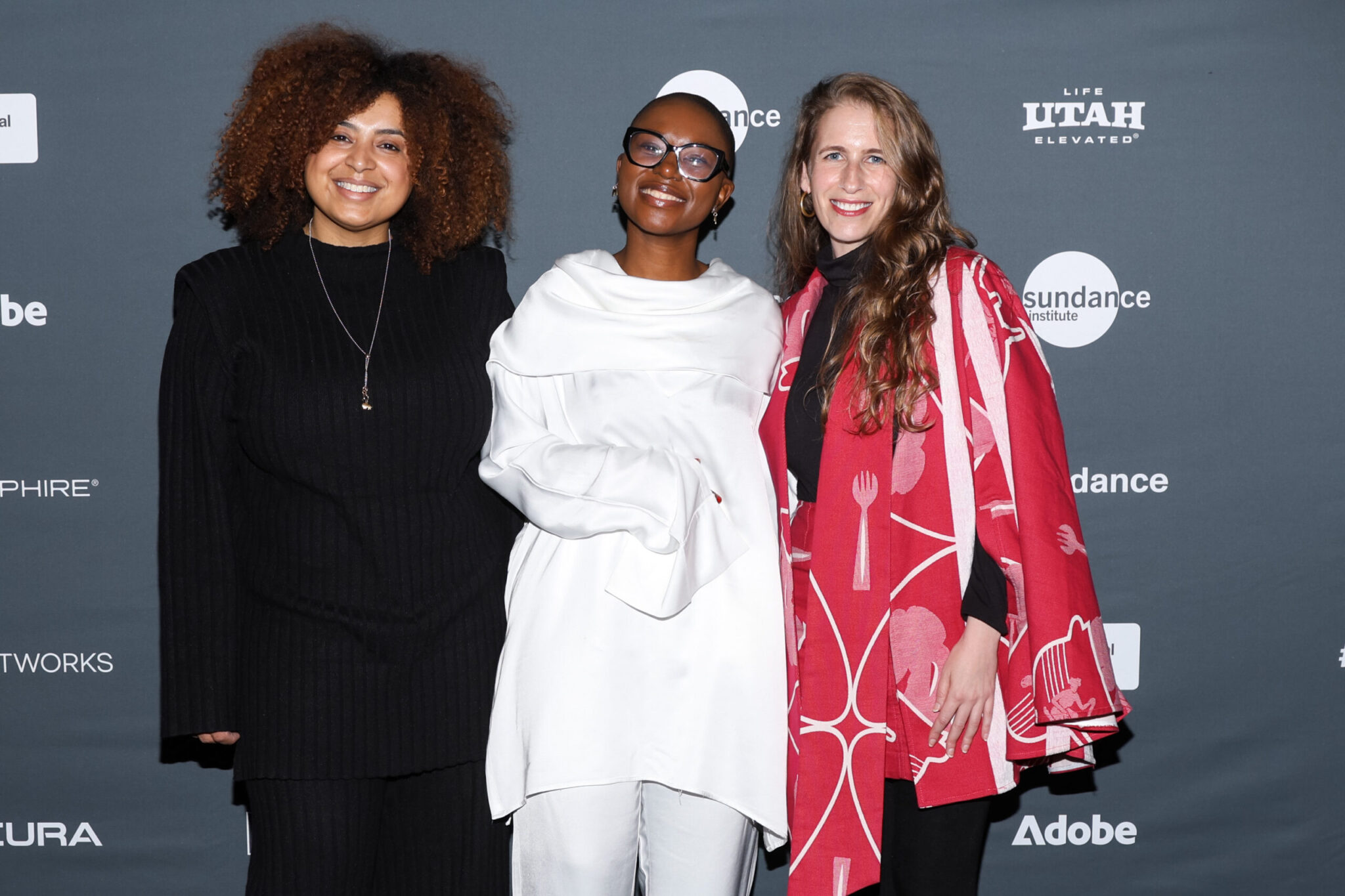 A woman dressed in all black, a woman dressed in all white, and a woman in a red dress, stand in front of a step and repeat at the 2023 Sundance Film Festival.