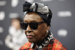 A close-up of a woman wearing a red plaid coat and a black headscarf and sunglasses.