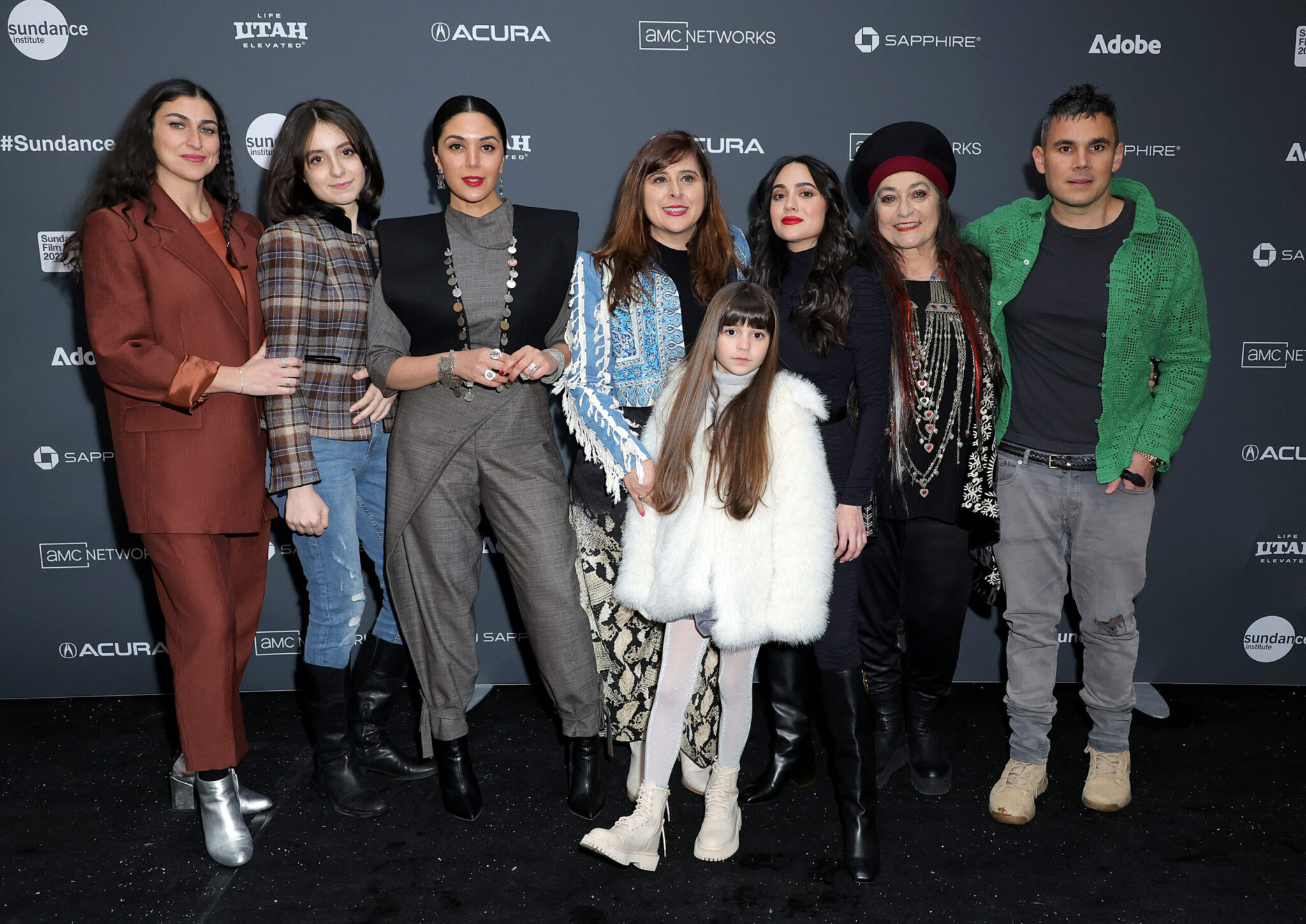 From left to right: A woman with long dark in a brown suit, a woman with medium-length dark hair and a checkered coat and jeans, a woman with black hair and a grey suit and black vest, a woman with light brown hair in a blue jacket, a little girl with long light brown hair in all white, a woman with long black hair in all black, a woman with long black hair and a black and red hat dressed in all black and wearing all-silver necklaces, and a man with black hair and a green jacket over an all-black outfit, stand in front of a step and repeat at the 2023 Sundance Film Festival.