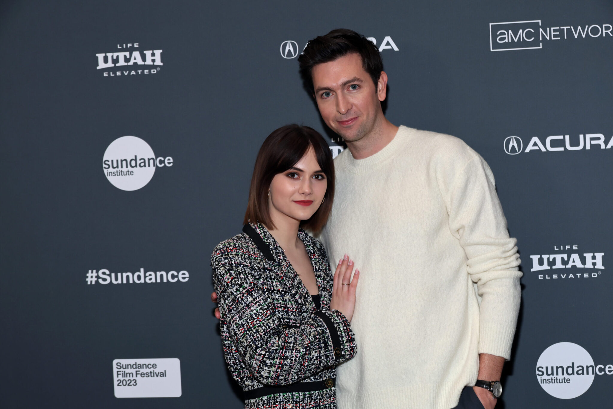 A woman with short brown hair and a blakc and white tweed coat stands with a tall brown-haired man in a white sweater.