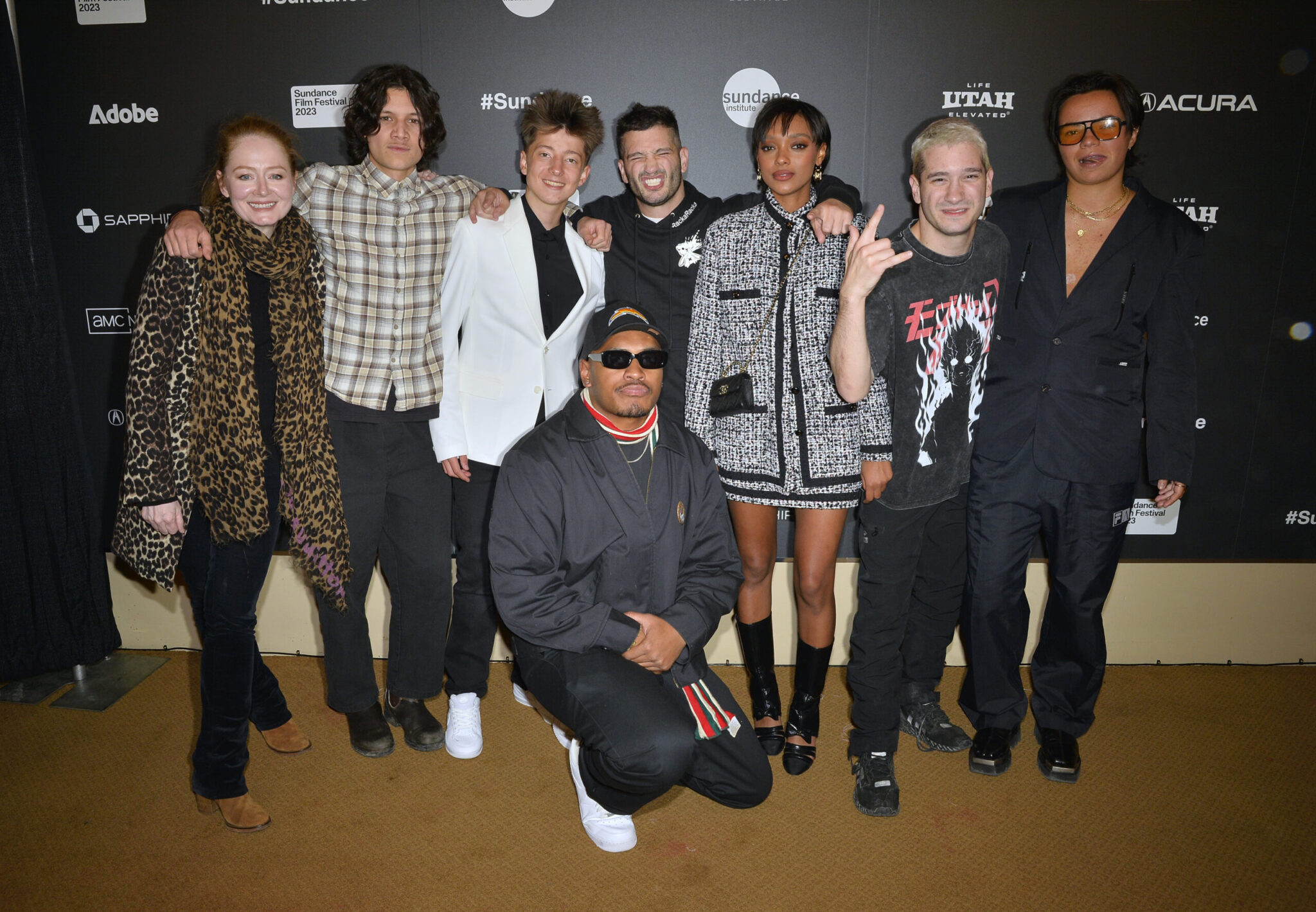 A woman with red hair and a leopard coat and scarf, a man with a plaid shirt and medium-length brown hair, a man in a white coat with spikey light brown hair, a man in a black hat and sunglasses dressed in all black kneeling in front, a man with blakc hair in all black, a woman with short black hair and a black and white tweed suit, a man with blonde hair and a black t-shirt, a man with black hair in an all-blakc suit, stand in front of a step and repeat at the 2023 Sundance Film Festival.