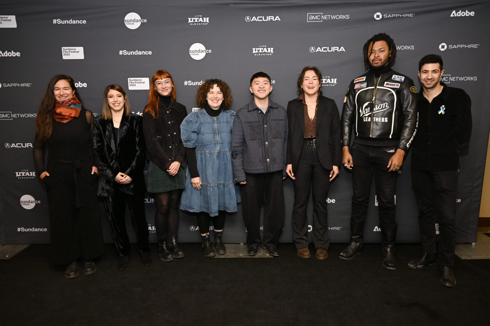 A woman with long brown hair and an orange scarf, a woman with short brown hair in all black, a woman with long red hair in a black coat, a women with brown curly hair in a denim dress, a man with short black hair and a charcoal jacket, a woman with brown hair and a black suit over a drown top, a man with black dreads in all black, a man with black hair in all black, all stand in front of a stand and repeat at the 2023 Sundance Film Festival.