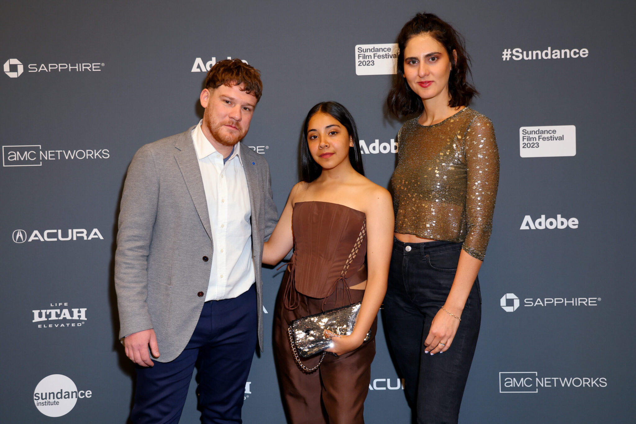 A bearded man with read hair, a white dress shirt, and grey blazer, and woman with long brown hair in a brown tube top and and brown pants, and a woman with short black hair, a sparkly long-sleeved blouse and black pants, all stand in front of a step and repeat at the 2023 Sundance Film Festival.