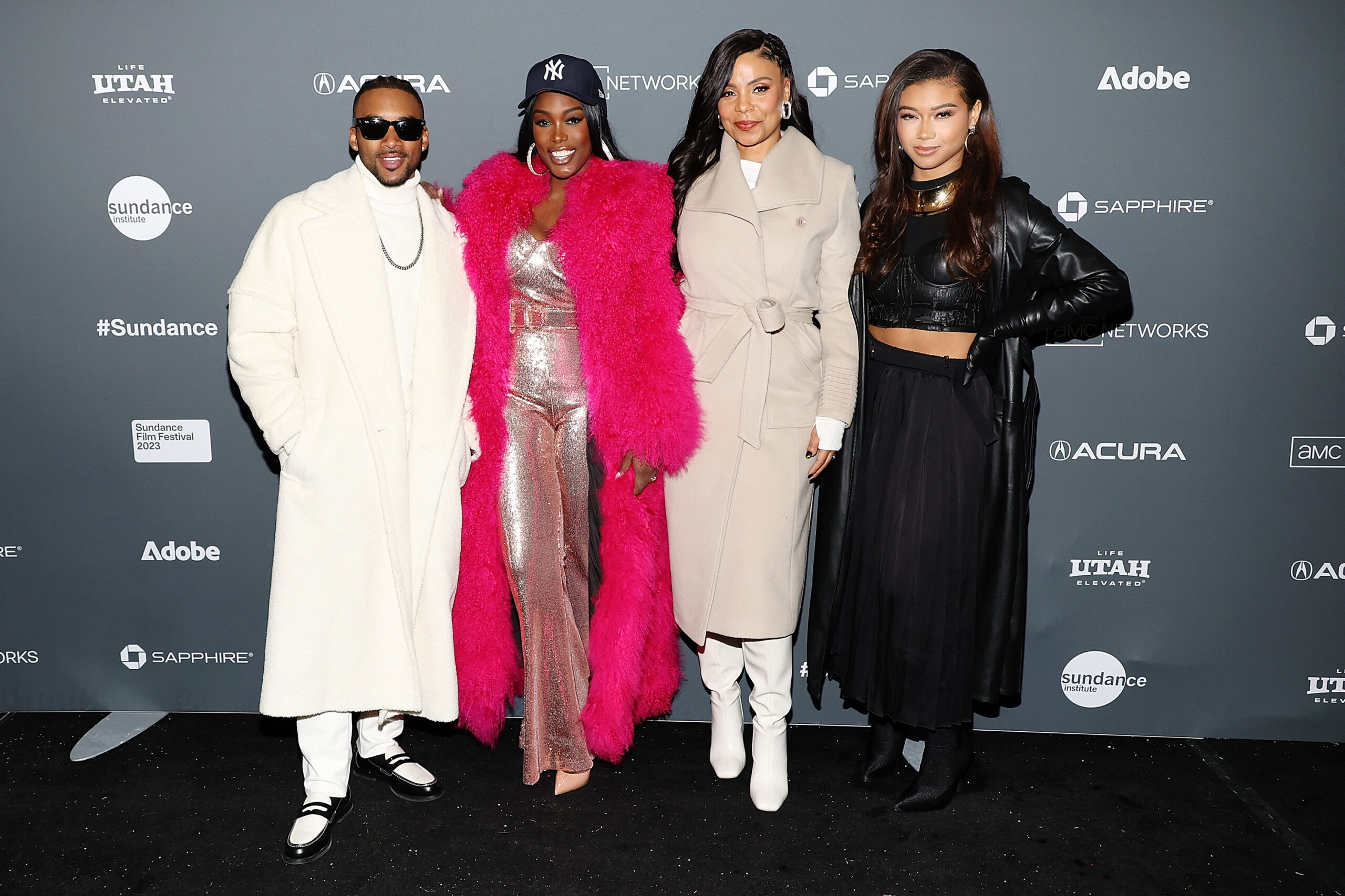 A man in all white, a woman in a long hot pink fur coat, a woman in all white, and a woman in all black stand in front of a step and repeat at the 2023 Sundance Film Festival