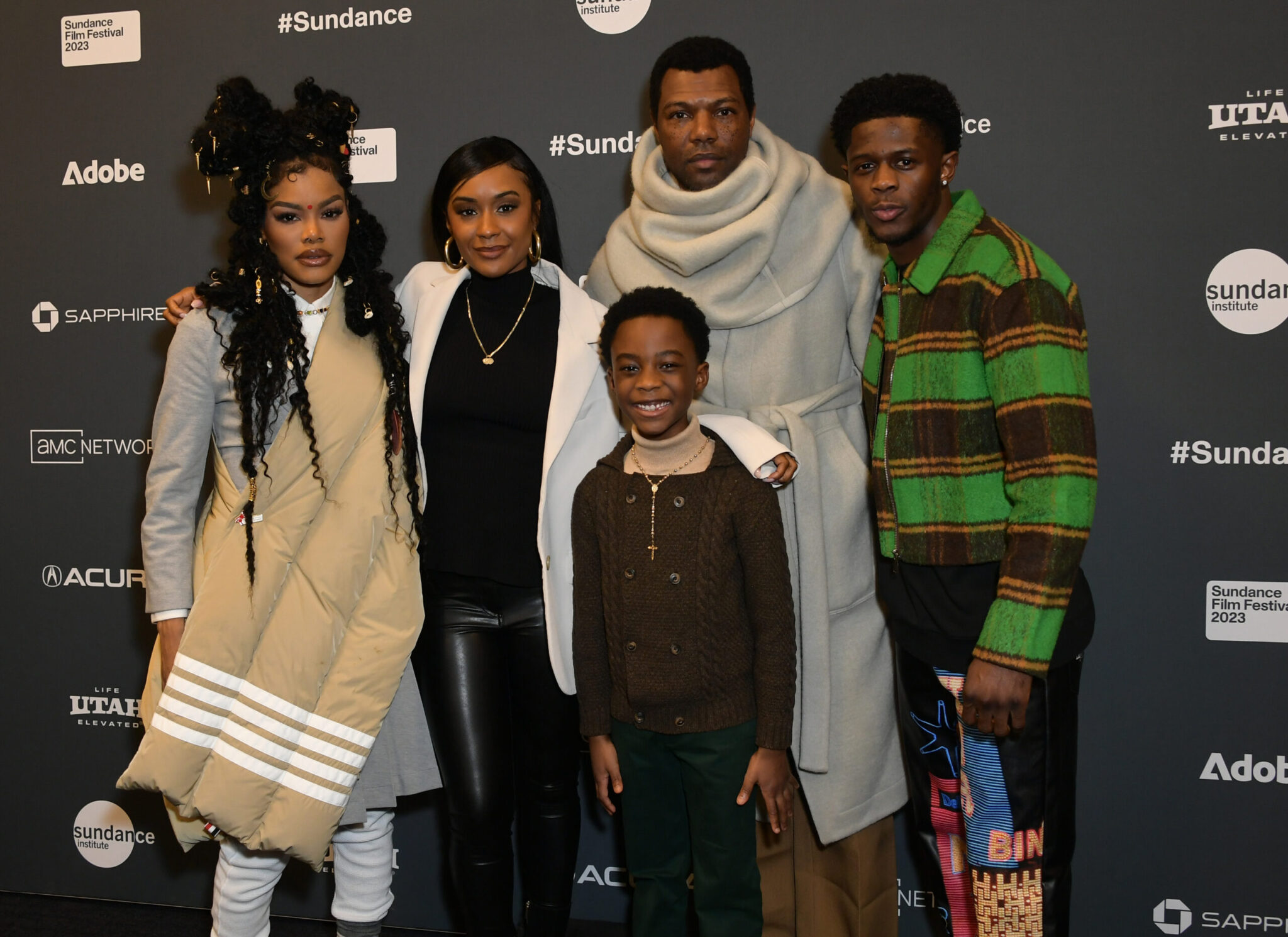 A woman in grey with a beige crossover, a woman in black with a black turleneck, a little boy in a brown sweater, a man with black hair in a grey poncho, and a man in a green and brown striped jacket, all stand in front of a step and repeat at the 2023 Sundance Film Festival