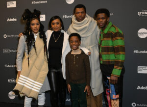 A woman in grey with a beige crossover, a woman in black with a black turleneck, a little boy in a brown sweater, a man with black hair in a grey poncho, and a man in a green and brown striped jacket, all stand in front of a step and repeat at the 2023 Sundance Film Festival