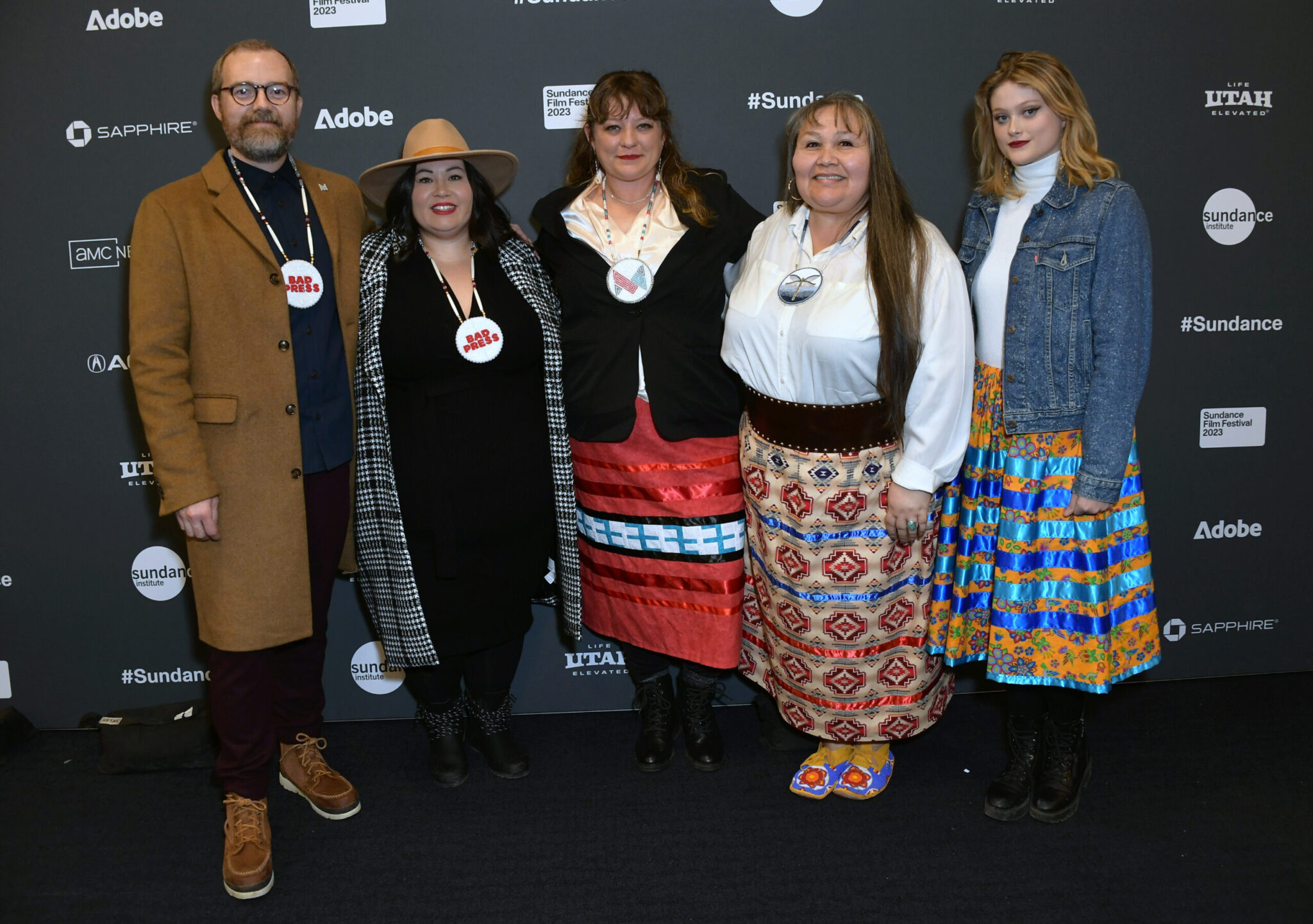 A bearded man with glasses and brown hair in a brown jacket over a blue shirt, a woman with black hair dressed in black with a black and white jacket and a brown hat, a woman with brown hair and a black blazer over a white blouse and red skirt, a woman with long brown hair in a white blouse, and a blonde woman in a jean jacket and a white shirt, all stand in front of a step and repeat at the 2023 Sundance Film Festival.