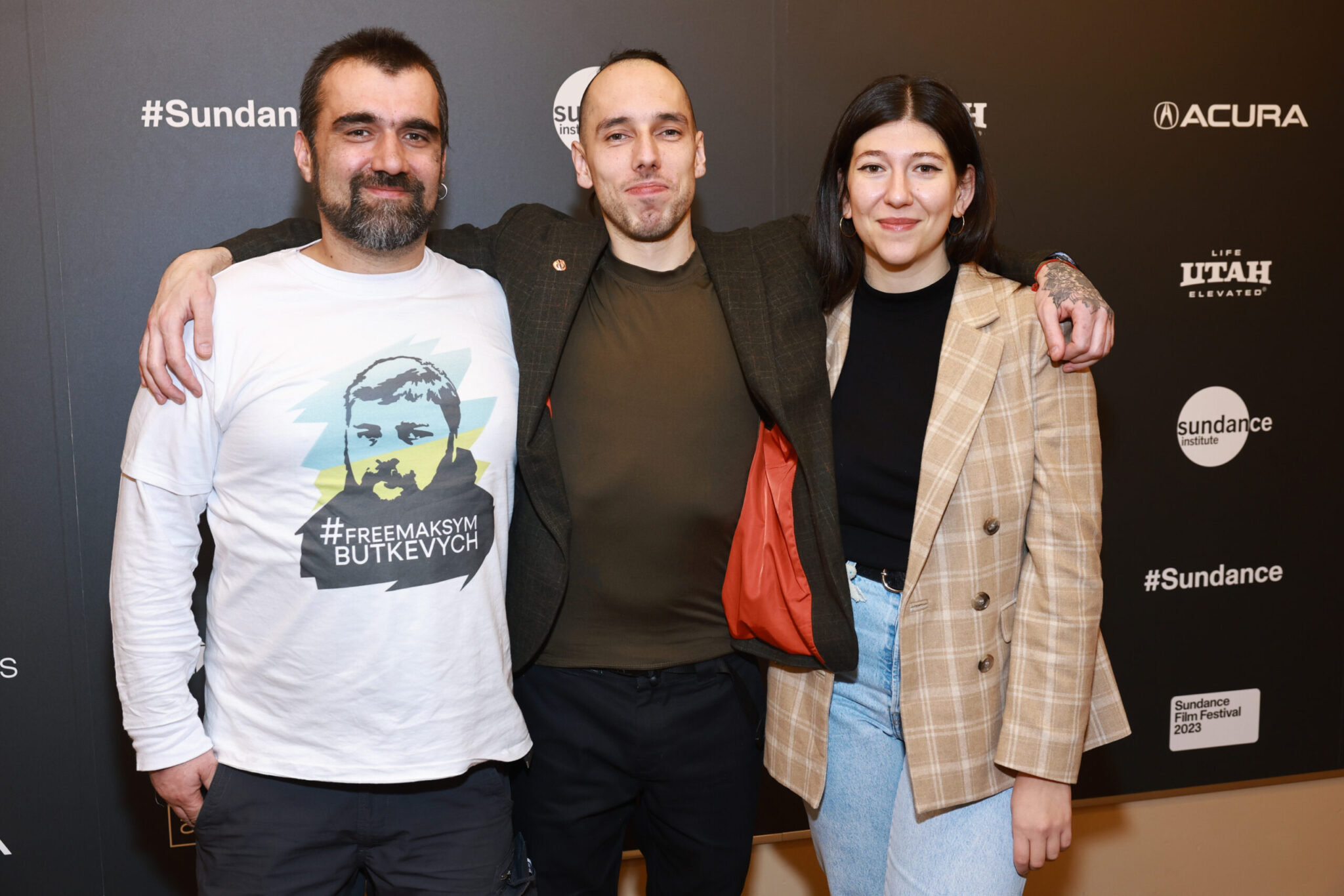 A man with a dark brown hair and beard in a white long-sleeved shirt, a man in a brown blazer with his arms around both the beard man and a woman in a tan blazer with short black hair, in front of a step and repeat at the 2023 Sundance Film Festival.