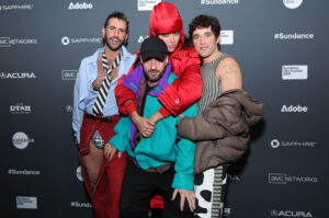 A man with spiked hair, a black and white tie, and a light blue dress shirt opened, a woman in all red hugs a man in a black hat and a 90s retro-style windbreaker, and a man in a striped outfit all stand in front of a step and repeat at the 2023 Sundance Film Festival.