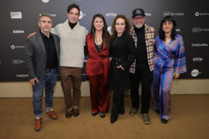 A man with grey hair and a grey blazer, a man with black hair and a tan sweater, a woman with long black hair in a red dress, a woman with brown curly hair in a long black dress, a beared man with a black hat in a checkered jacket and glasses, and a black-haired woman in a blue dress, all stand in front of a step-and-repeat at the 2023 Sundance Film Festival.