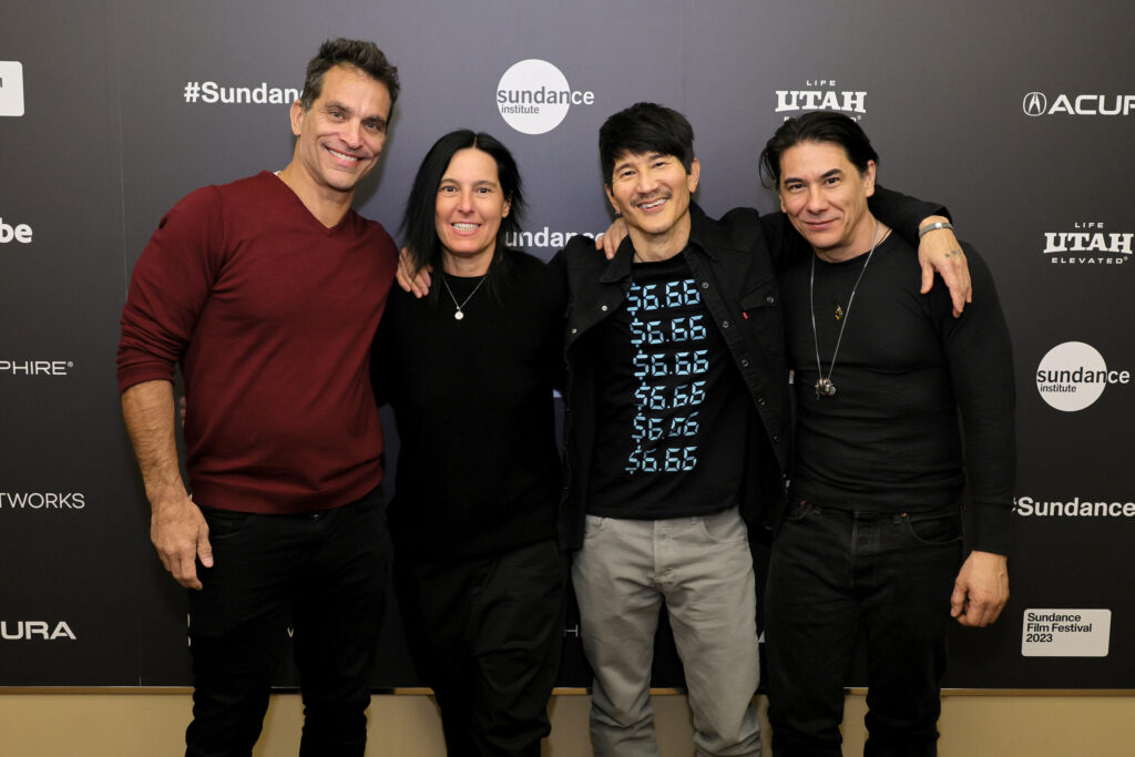 A tall man with dark hair and a red shirt, a woman with medium-length black hair dressed in black, a taller man with short black hair and a blck shirt with writing on it, and a medium-height man with his hair tied back dressed in black.