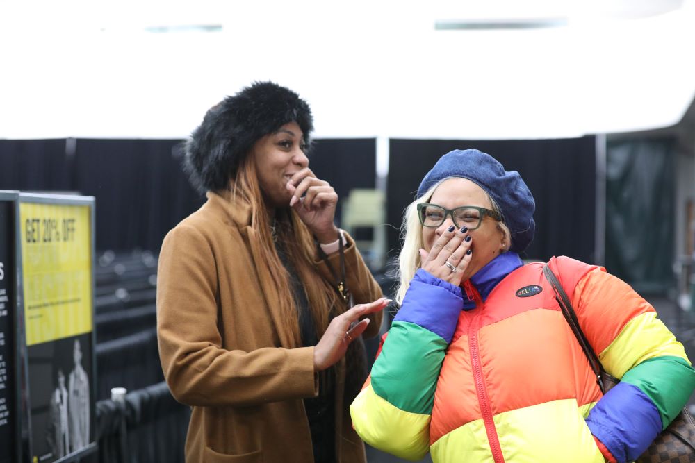 Two women dressed in winter coats and hats share a giggle on a street.
