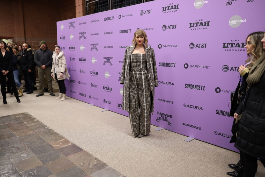 Woman with long blond hair and red lipstick poses in front of the Sundance Institute backdrop, dressed in a plaid jumpuit and matching long coat. Observers dressed in winter outerwear watch from two sides