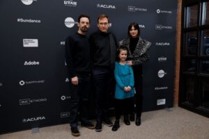 From left to right: A man dressed in black with a beard and black hair, a man with brown hair dressed in all black, a little girl with brown hair in a blue dress and black pants, a woman with black hair dressed in all black , all stand in front of a step and repeat at the 2023 Sundance Film Festival.