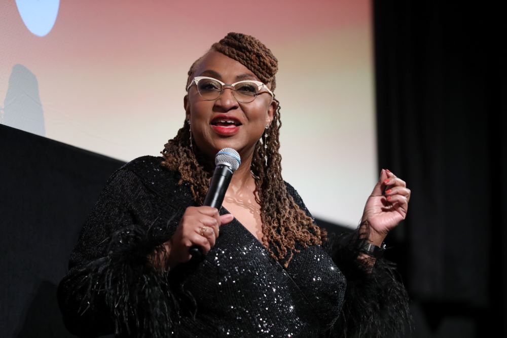 A dark-skinned woman with braided and curly long hair, wearing a sparkly black outfit, speaks into a microphone.