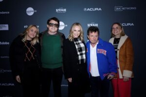 From left to right: A blonde woman dressed in black, a man with dark brown hair in a green sweater and black coat with black sunglasses, a blonde woman with a black and white checkered scarf, a man with black hair and a blue jacket over a white shirt, and a blonde woman with a brown jacket and red pants, all stand in front of a step and repeat at the 2023 Sundance Film Festival.